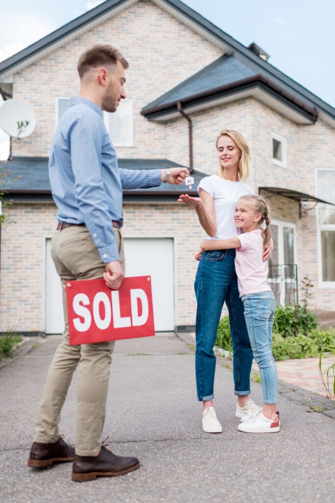 real estate agent with sold sign giving key to young woman with daughter in front of new house